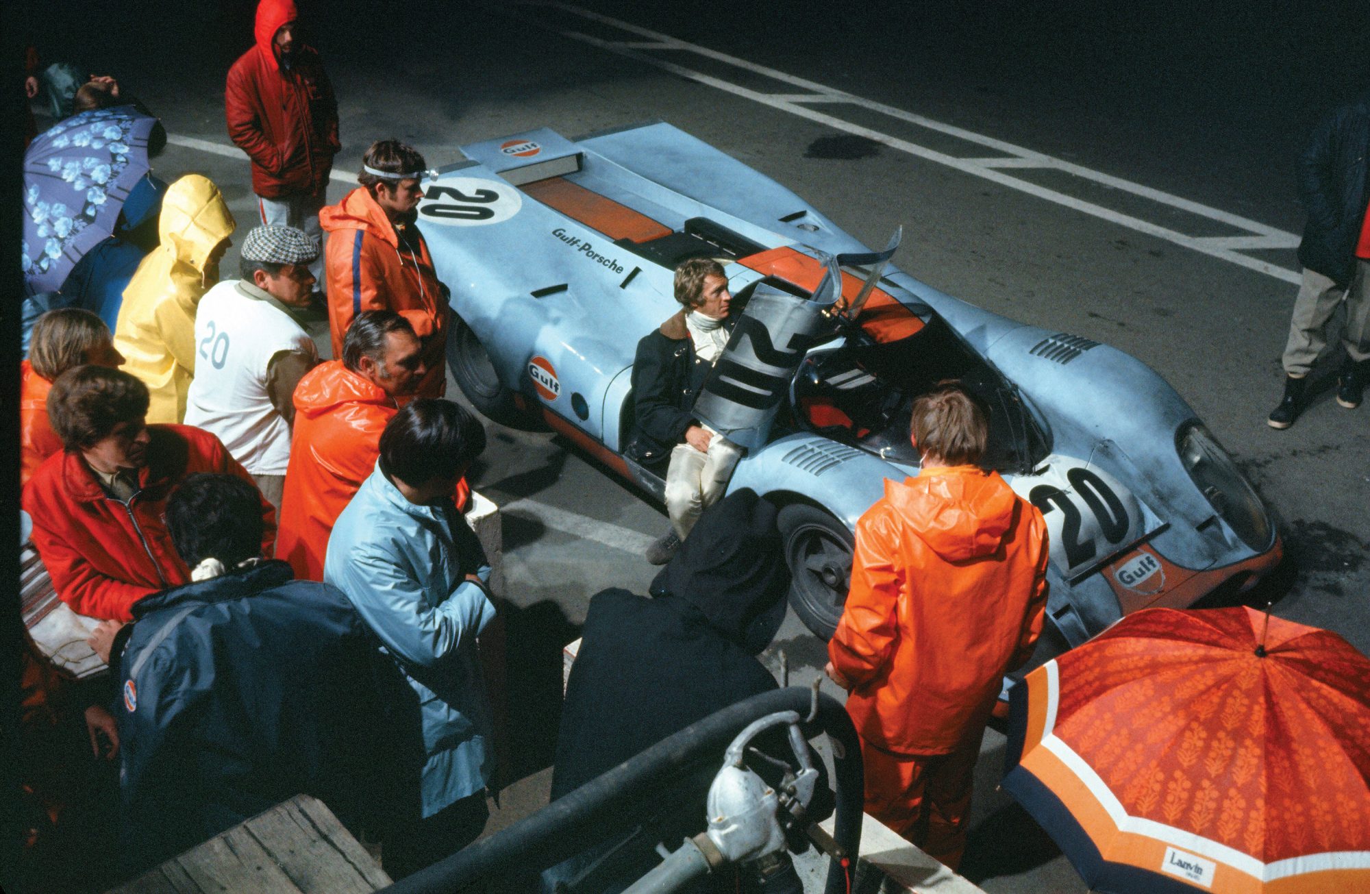 Steve McQueen sitting in a Porsche 917KH with number 20 Gulf Oil livery, surrounded by crew of the film